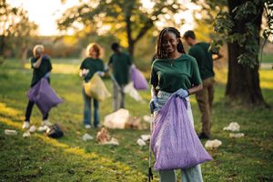 Group of volunteers in green shirts clean a park, picking up trash outdoors on a sunny day. Smiling, they work together, showing care for the environment and promoting sustainability